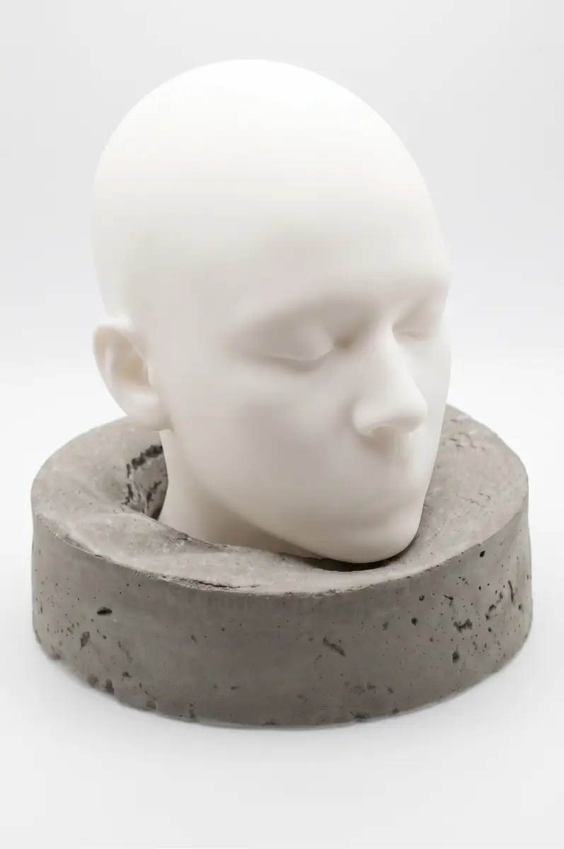 Escultura O Peso do Silêncio vista de semi-perfil. Cabeça humana branca com olhos fechados inserida em uma base circular cinza de concreto, destacando o encaixe da peça. Fundo branco.