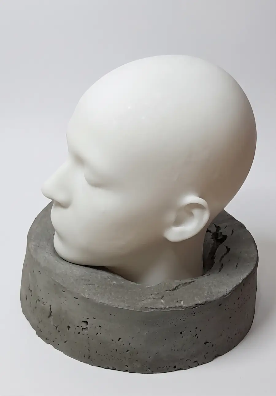 Vista de perfil esquerdo da escultura O Peso do Silêncio, do artista Moreira. Cabeça humana branca com olhos fechados e feições suaves inserida em uma base cilíndrica de concreto cinza poroso. Fundo branco.